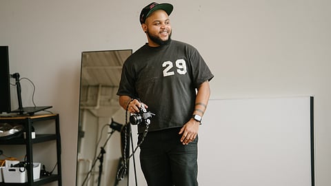 A Black trans man with a beard and black baseball cap holds a camera.