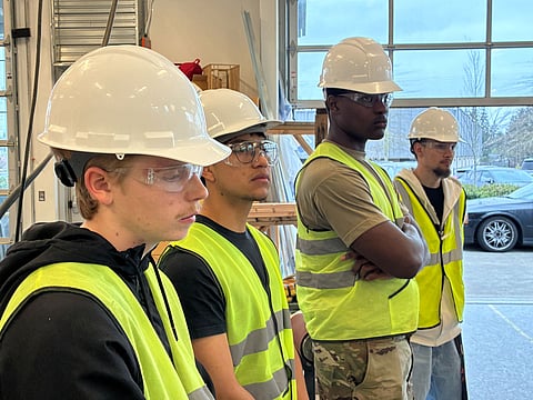 Four high school seniors stand side by side intently looking at something off camera. They're wearing high-visibility vests and hard hats.