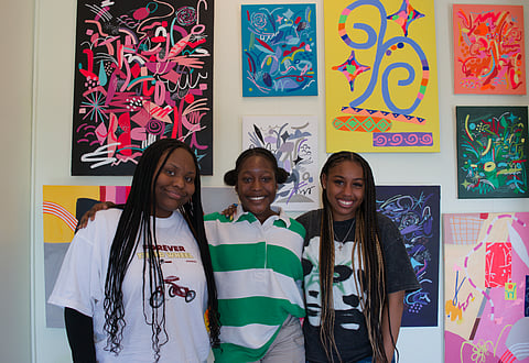 Three young Black women stand shoulder to shoulder in front of a wall of colorful paintings.
