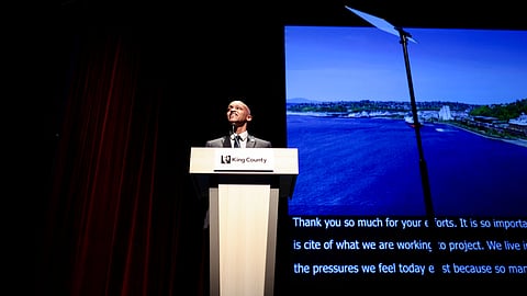 Girmay Zahilay speaks at a podium during a public address, with a waterfront image displayed on a large screen behind him.