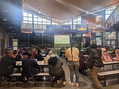 Dozens of community members sit at cafeteria tables facing a presentation screen.