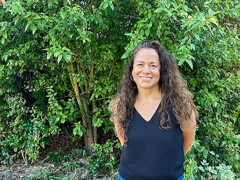Laura van Dernoot Lipsky stands with her hands behind her back in front of foliage.