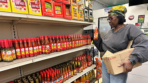 Sophia Ellis stocks chili sauce in her shop, Just Like Home Caribbean Market and Kitchen. She's holding a cardboard box against her hip and pulling out hot sauces to put on the shelves.