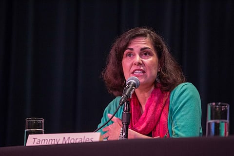 Close-up of a woman (Tammy Morales) with short, wavy dark hair at a microphone.