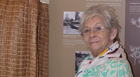 A woman with short gray hair and glasses stands beside a display in a museum. She is wearing a patterned green top and a colorful scarf. Behind her are informational panels and historical photos.