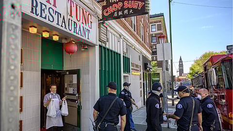 A restaurant worker, wearing an apron and holding two plastic bags, stands at the entrance of Tai Tung Restaurant. Several firefighters in uniform and face masks gather nearby on the sidewalk next to a fire truck. The restaurant sign reads "Chop Suey."