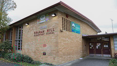 The exterior of Graham Hill Elementary School, a brick building with large windows. The school name is prominently displayed in red lettering on the wall. A banner hangs on the building, recognizing it as a 2017-18 Washington State Recognized School. The entrance features two red doors, and there are small trees and bushes surrounding the building.