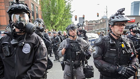 A group of police officers in tactical gear, helmets, and gas masks stand in formation during a protest in Seattle. Some officers hold batons or other equipment, and others wear body cameras. The background shows city buildings, trees, and traffic lights. The officers appear focused and alert.
