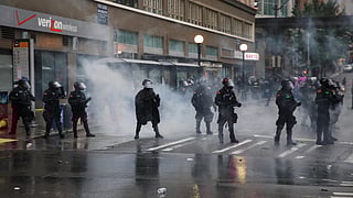 A group of riot police officers in black uniforms and helmets with face shields stand in formation on a wet city street filled with tear gas or smoke. Some officers hold batons or firearms.