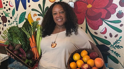 Central Cafe and Juice Bar owner Bridgette Johnson poses with fresh produce in front of a welcoming mural in her cafe. Photo courtesy of Bridgette Johnson.