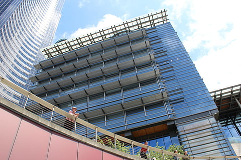 Modern glass building with a grid-like sunshade structure on its upper levels, viewed from below against a blue sky with clouds. A man in a plaid shirt and cap leans on a railing in the foreground, while other individuals are partially visible along the walkway.