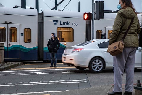 Trains and cars whiz by as people wait to cross Martin Luther King Jr. Way at the Othello Station. Pedestrians, cyclists, and motorists navigate these crossings along the 4.5-mile light rail stretch through the South End. (Photo: Lizz Giordano)