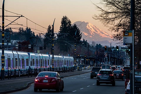 Martin Luther King Jr. Way South with cars and a light rail train head down the street. Mt. Rainier lit up by a sunset looms in the background.