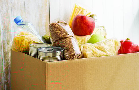 A cardboard box filled with assorted food items, including canned goods, a water bottle, packaged pasta, a bag of buckwheat, a red apple, a green pear, a bottle of cooking oil, and a red bell pepper, placed against a rustic wooden background.