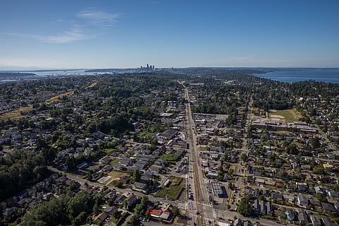 Aerial photo of Rainier Valley in Seattle, showing dense residential neighborhoods, Rainier Avenue, and distant view of downtown skyline and Puget Sound under a clear blue sky.