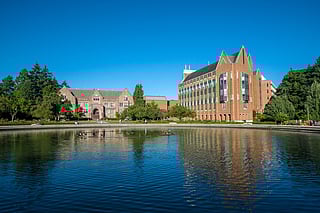 Brick academic buildings at the University of Washington reflected in a calm pond, with clear blue skies above and trees surrounding the scene.