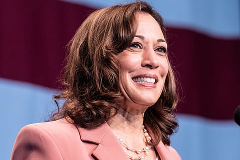 A close-up photo of Kamala Harris smiling while speaking at an event. She is wearing a pink blazer and a pearl necklace, with her wavy brown hair framing her face.