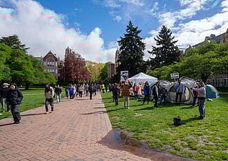 A peaceful protest on a university campus with participants and tents on a grassy area, with campus buildings and students walking by in the background under a partly cloudy sky.