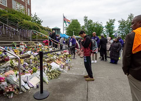 People gather around a memorial on Garfield High School's steps filled with flowers, candles, notes, and a teddy bear. One young man stands in front, head bowed and hands clasped. Others, wearing jackets and backpacks, observe quietly. Flags fly in the background, and the atmosphere is solemn and respectful.