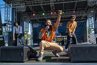 Three performers on an outdoor stage engage the audience energetically. The central figure, in an orange dashiki, kneels with a raised fist while holding a microphone. The other two wear stylish outfits and hold microphones. The stage features banners for