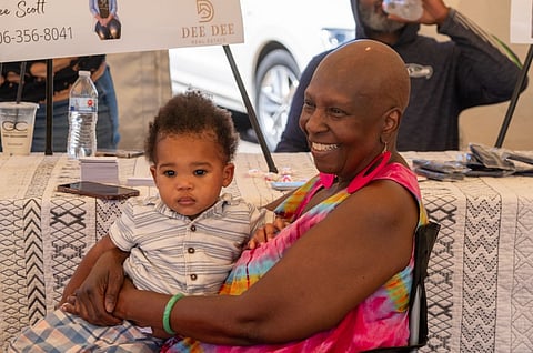 A smiling elderly woman with a shaved head and vibrant pink earrings holds a young child on her lap. They are at a table covered with a patterned cloth, with a phone and water bottles nearby. A