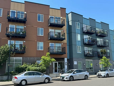A modern apartment building with two sections, one in brown and the other in blue-gray, featuring multiple balconies with potted plants. Three cars are parked in front of the building on a sunny day, with a clear blue sky above. The entrance to the building is visible, and small trees and shrubs line the sidewalk.