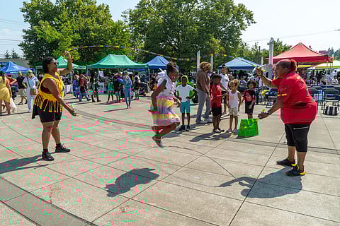 Black-presenting girl in a rainbow dress jumping rope at an outdoor community event, with two Black, femme-presenting adults turning the rope and families gathered under colorful tents in the background.