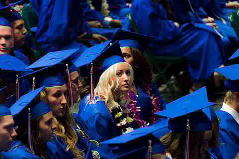 A crowd of students in blue graduation caps and gowns, with the photo centered on a blonde student in the center.