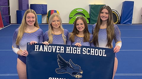 A group of 4 female Hanover high school gymnasts hold their schools blue school flag that says Hanover High School Gymnastics on it with a blue hawk on it as their mascot