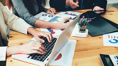 Three people sit at a wooden table with spreadsheets and charts laid out around them looking at a computer and a tablet.