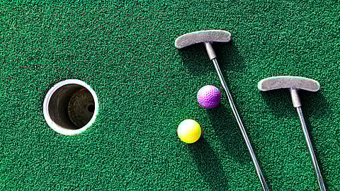 Putting green turf with two putters, and yellow and pink balls next to a golf hole.