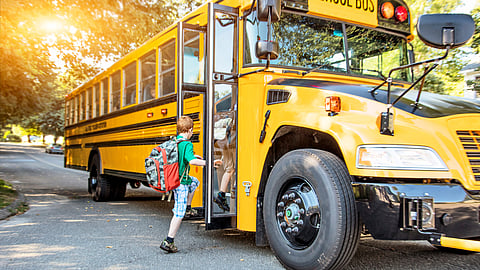 Red headed child wearing a green shirt blue shorts with a red backpack gets on a yellow school bus with the sun shining through the trees in the background