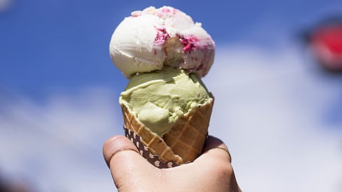 a hand holding two scoops of ice cream in a cone against a blue sky with clouds