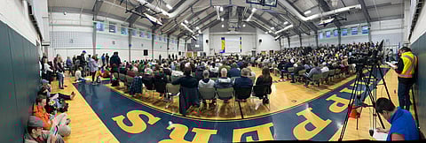gymnasium is filled with folding chairs and people for a town meeting