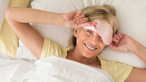 a white woman with blonde hair lays in bed with an eye mask on looking up