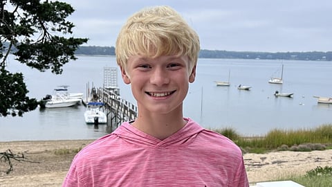 a young blonde haired man stands in front of the harbor with sailboats in the background. he is wearing a pink hoodie and smiling.
