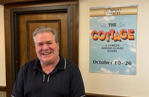A man posing in front of a poster for the production of "The Cottage"