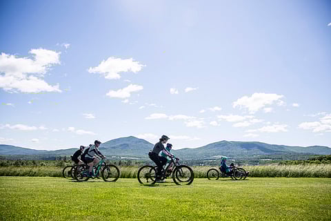 a group of women mountain bikers ride across a green field with mountains in the back and a blue sky dotted with fluffy white clouds above