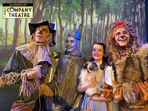 The scarecrow, tin man, Dorothy, Toto, and Lion stand in costume smiling with trees behind them