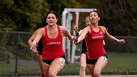 two hingham high school tracker runners wearing red jerseys pass a baton on the track