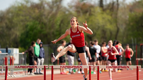 a student wearing a red jersey and black shorts leaps over hurdles on a track