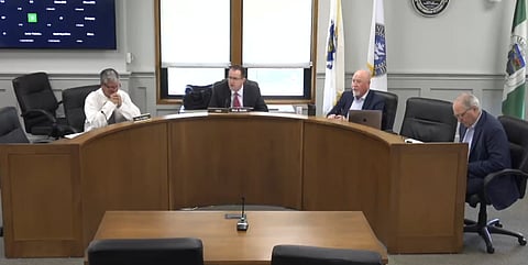 4 white men sit at a curved desk in a meeting room