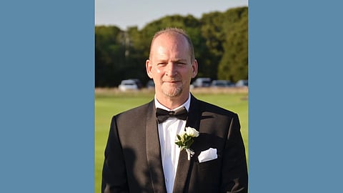 smiling middle aged white man with a light blonde beard and receding hairline in a black tuxedo with a white flower on the lapel