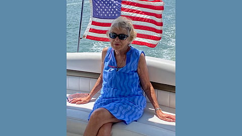 photo of an older white woman with white hair and dark sunglasses sitting on a boat on the water with an american flag waving behind her