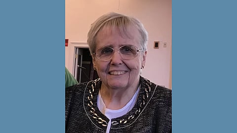 photo of older white woman with gold metal rim glasses smiling into the camera and wearing a black boucle jacket with gold chain trim