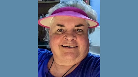 Image of an older white woman with white hair wearing a blue shirt and pink visor