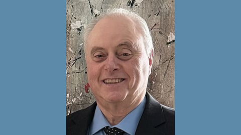 Older white man wearing a black suit with a light blue oxford and black dot tie smiles at the camera