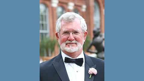 Older white man with white hair and beard and wire rimmed glasses wearing a tuxedo with a light pink carnation on the lapel smiles at the camera