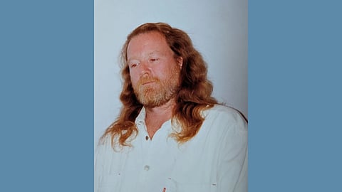 Photo of a middle aged white man with long dark blonde wavy hair and a beard wearing a long sleeve white button up shirt and looking down