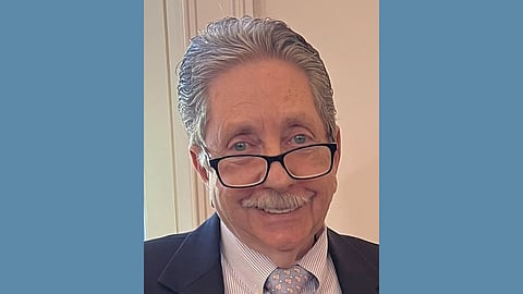 An older white man with silver hair and mustache wearing glasses low on his nose smiles warmly